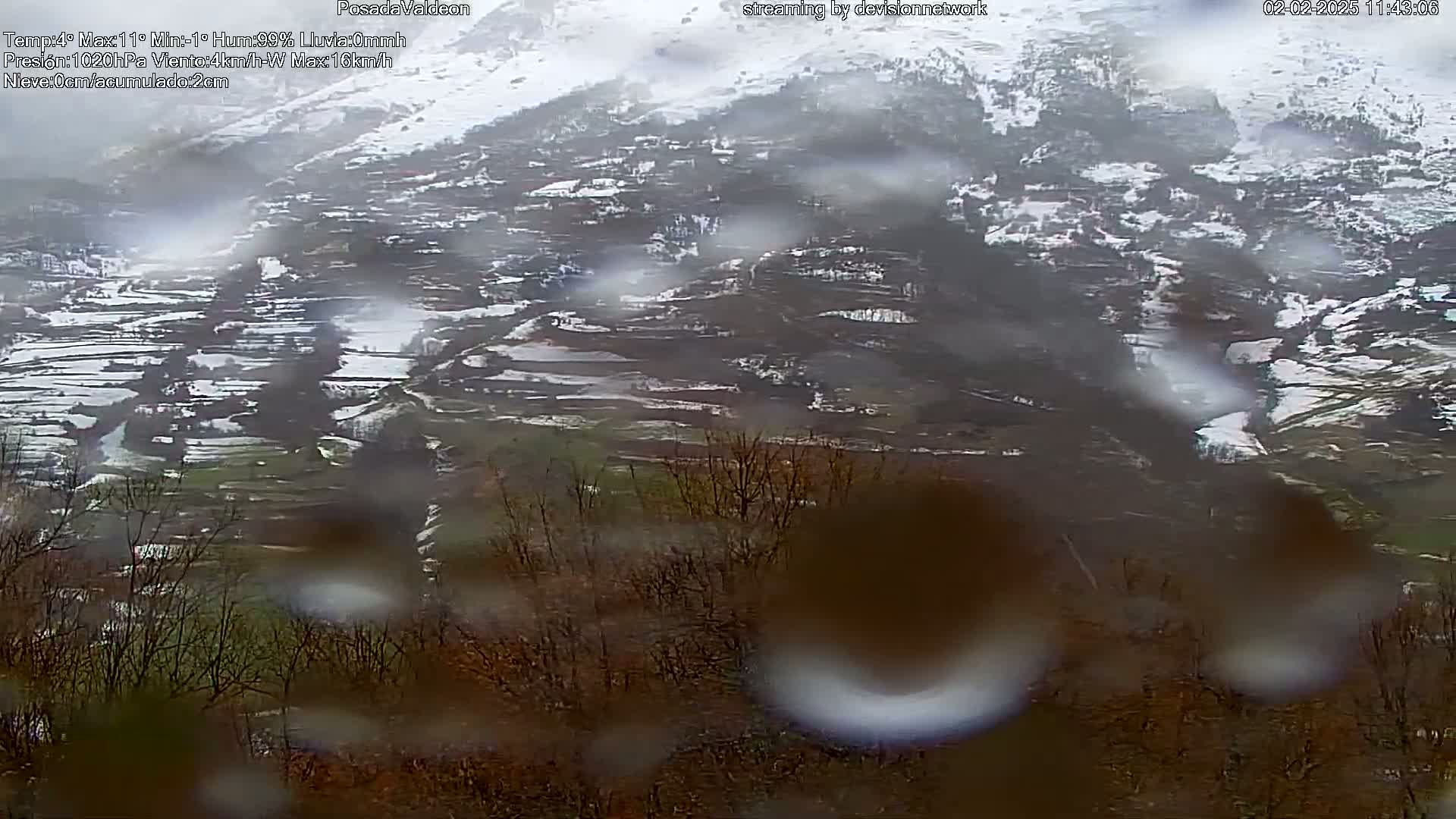 A rain-covered view of a snow-dusted valley with terraced fields and sparse trees.