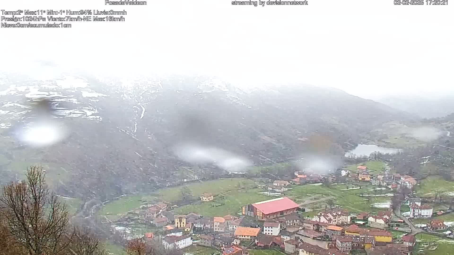 A small village nestled in a valley, surrounded by snow-dusted mountains, under a hazy, rainy sky.