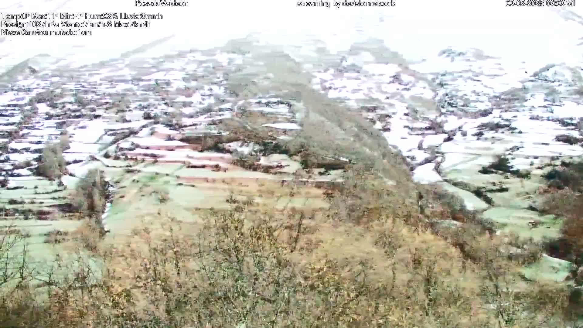 A snow-covered village nestled in a valley, viewed from a hillside, with light snow falling.