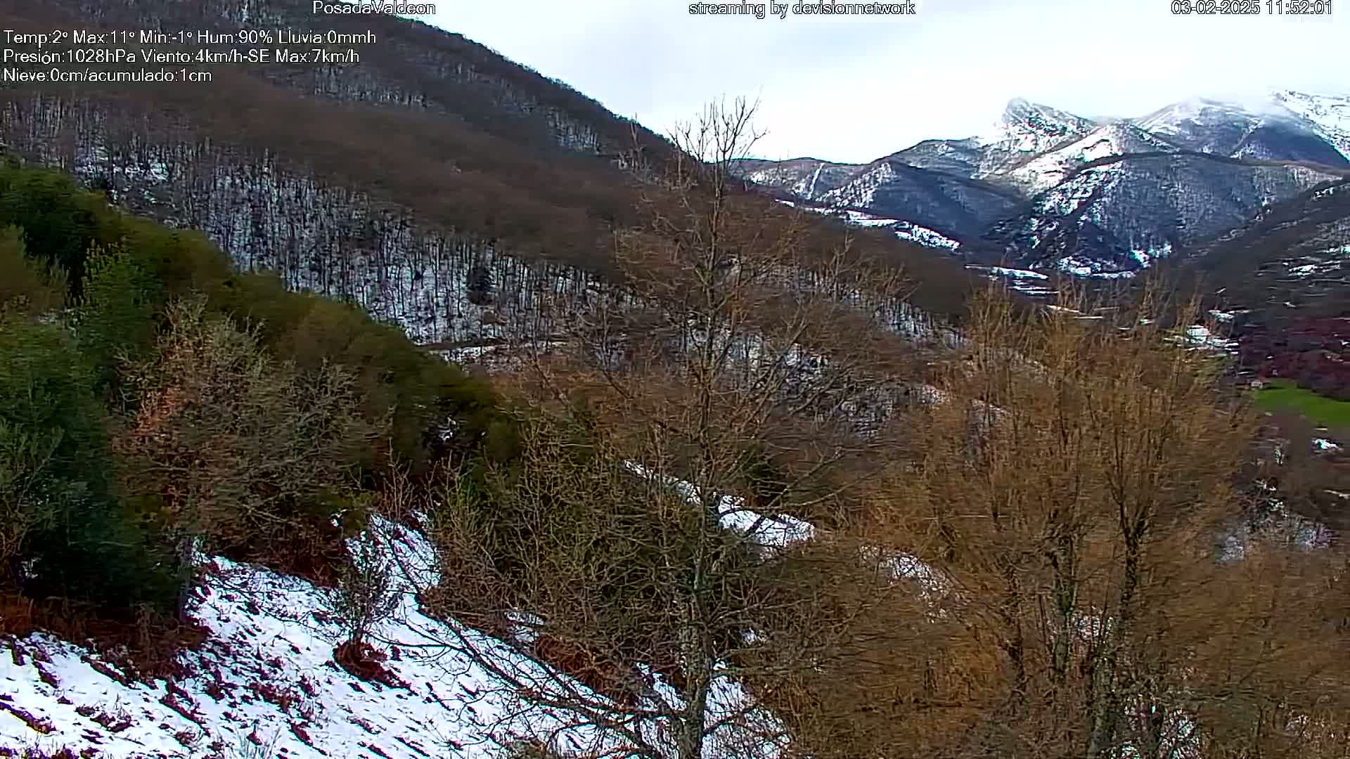 A snow-dusted mountain valley with bare, brown trees and patches of snow on the ground, under an overcast sky.