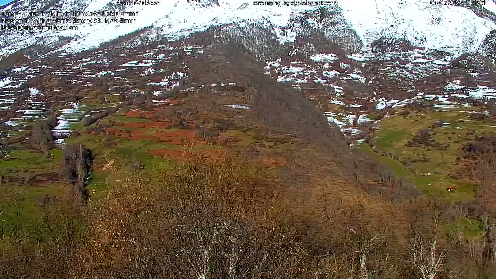 A snow-dusted mountain valley reveals a landscape of patchy green and brown fields, interspersed with leafless trees and a small stream, under a partly cloudy sky.