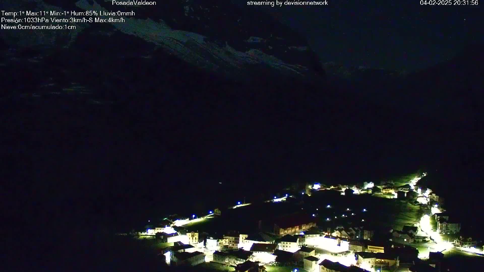 A nighttime, aerial view of a small, illuminated village nestled at the base of a dark mountain under a clear, dark sky.