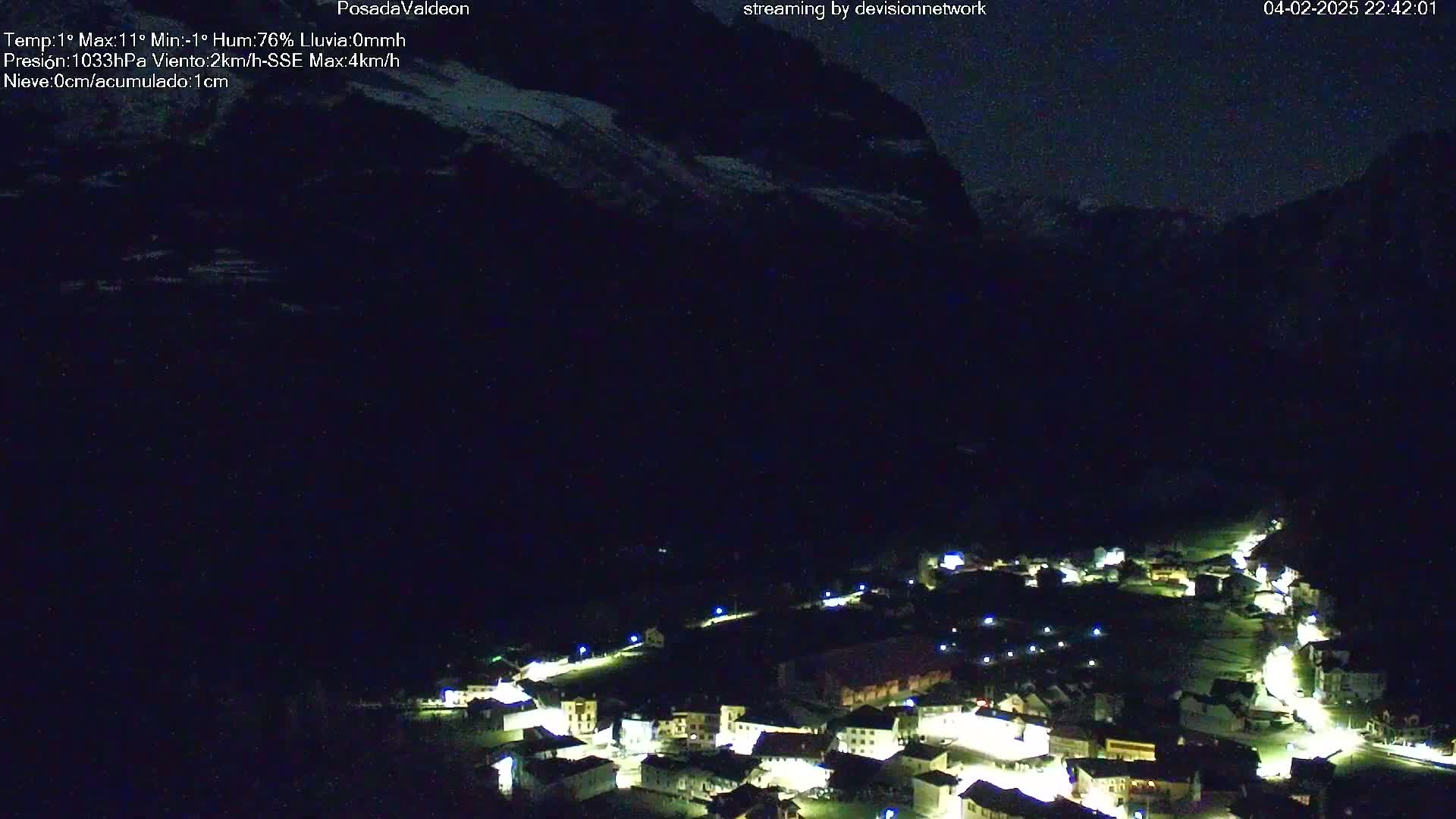 A nighttime, aerial view of a small, illuminated village nestled in a valley, with dark mountains and a hint of snow in the background under a dark sky.