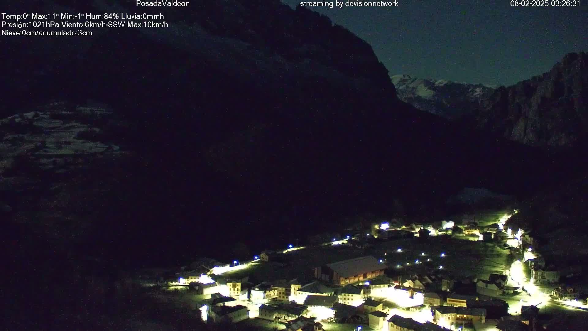 A small, illuminated village nestled in a dark valley below a large, snow-capped mountain under a night sky.