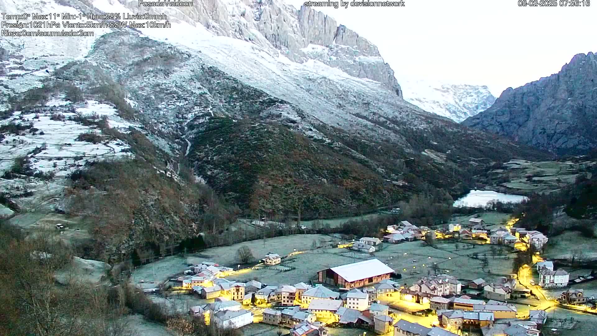 A small, illuminated village nestled in a frost-covered valley between snow-dusted mountains under a pale sky.