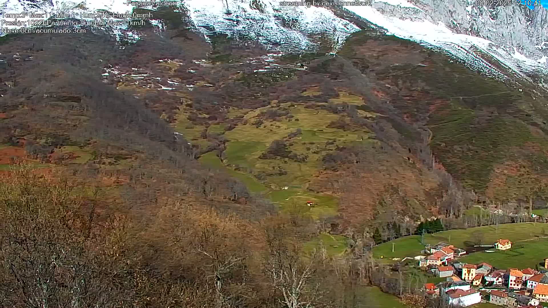 A snow-dusted mountain overlooks a valley with a small village nestled amongst brown and green fields.
