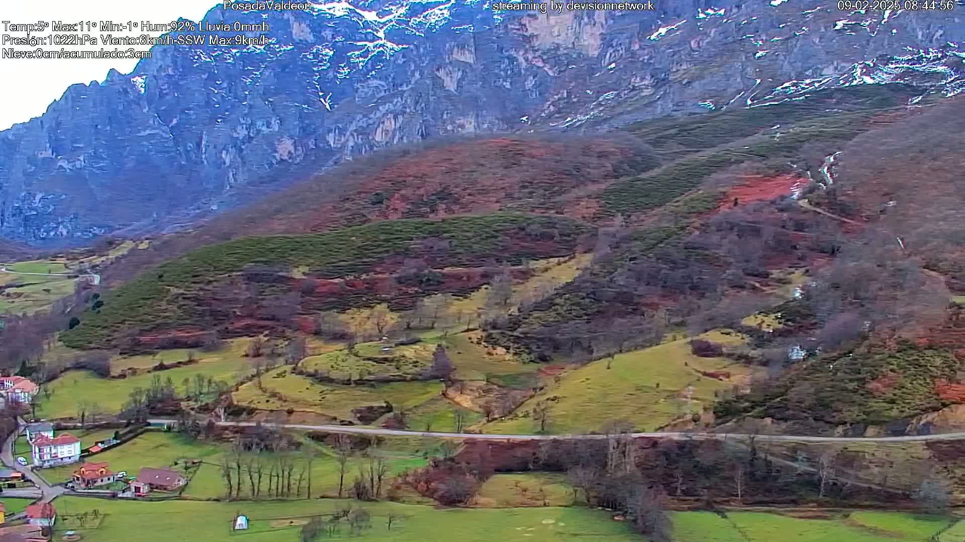 A picturesque valley with a road winding through green and brown fields, backed by a steep, snow-dusted mountain.