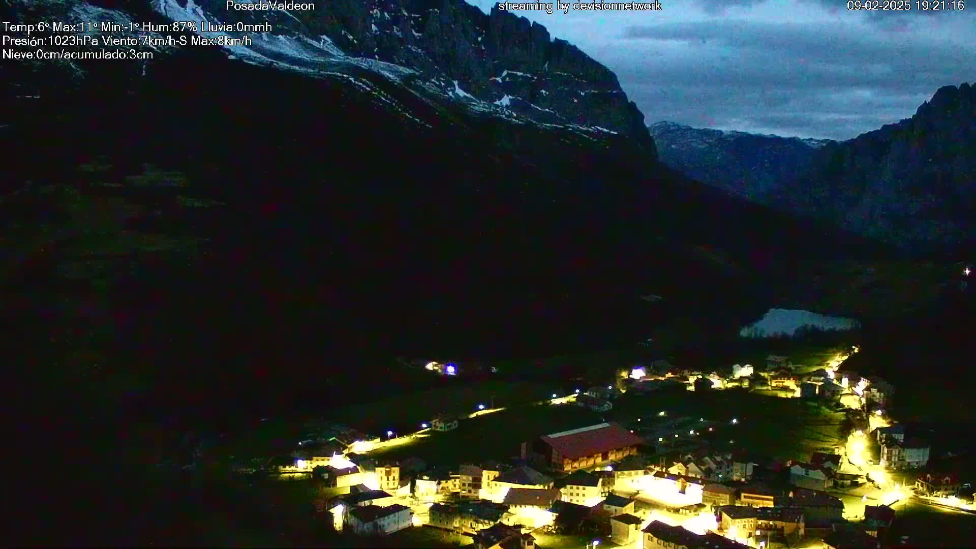A small, illuminated village nestled in a dark valley beneath a partly cloudy night sky and towering mountains.