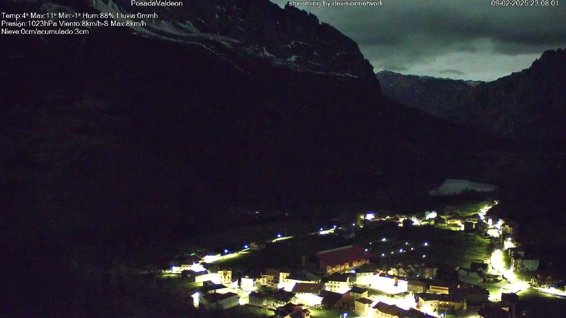 A nighttime view of a small village nestled in a valley beneath a dark, cloudy sky and a mountain range.