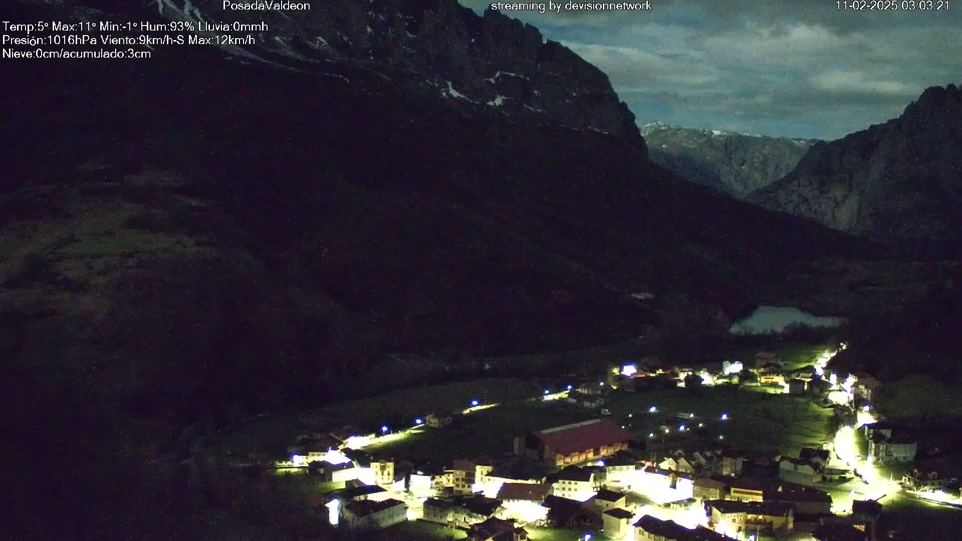 A small village nestled in a valley between dark mountains is illuminated at night under a cloudy sky.