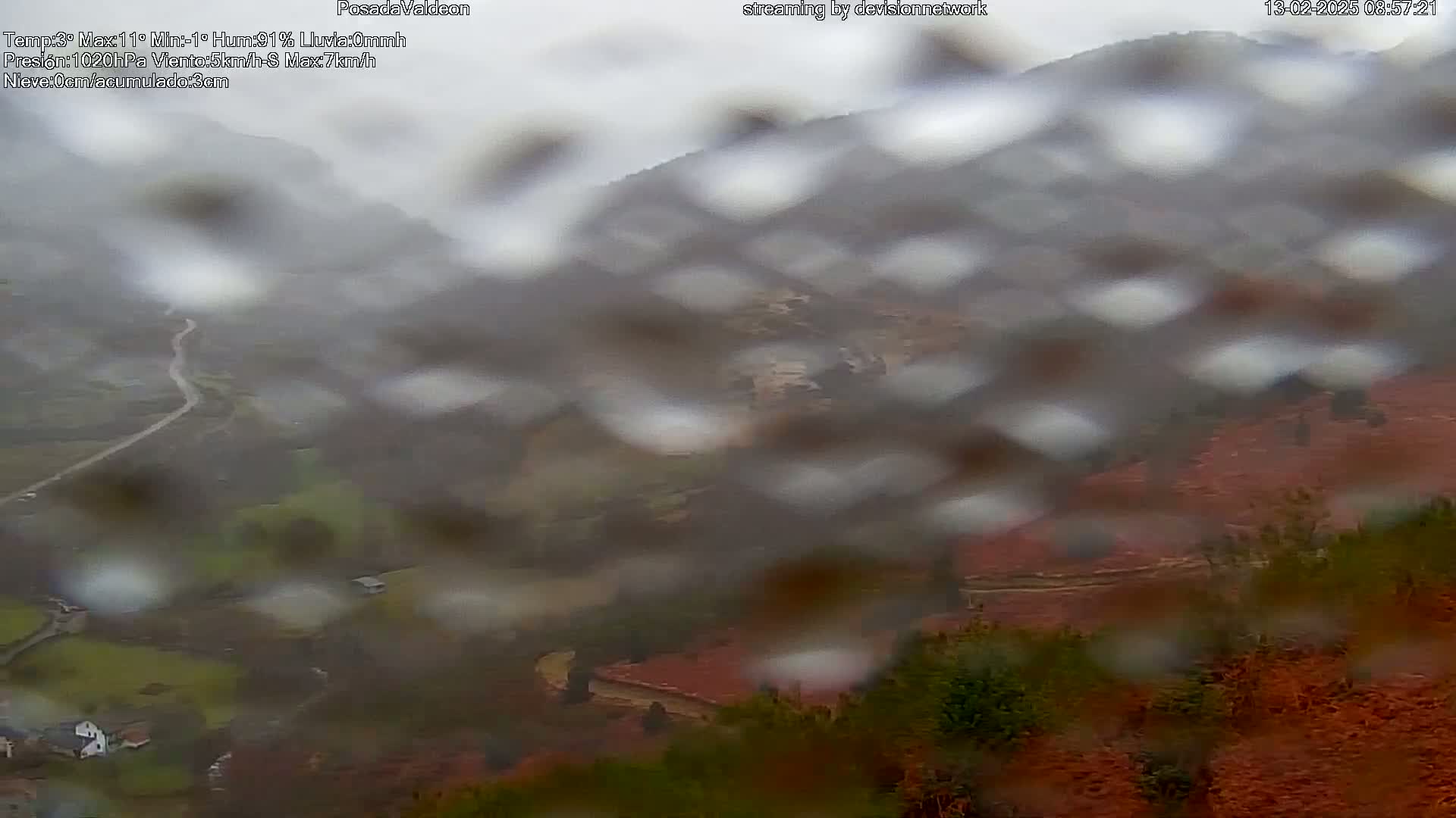 A rain-streaked view shows a winding road through a hilly landscape with patches of reddish-brown vegetation.