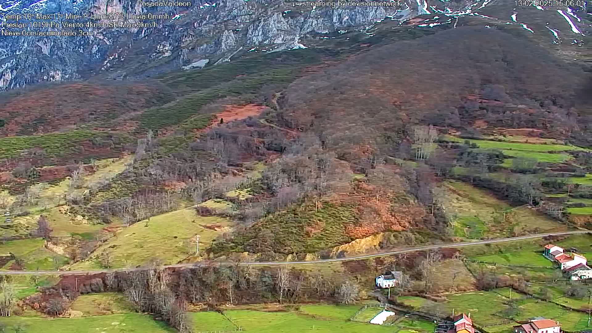 A high-angle view of a rural valley, with a road winding through fields and sparse deciduous trees, under overcast conditions and a mountain range capped with snow in the background.