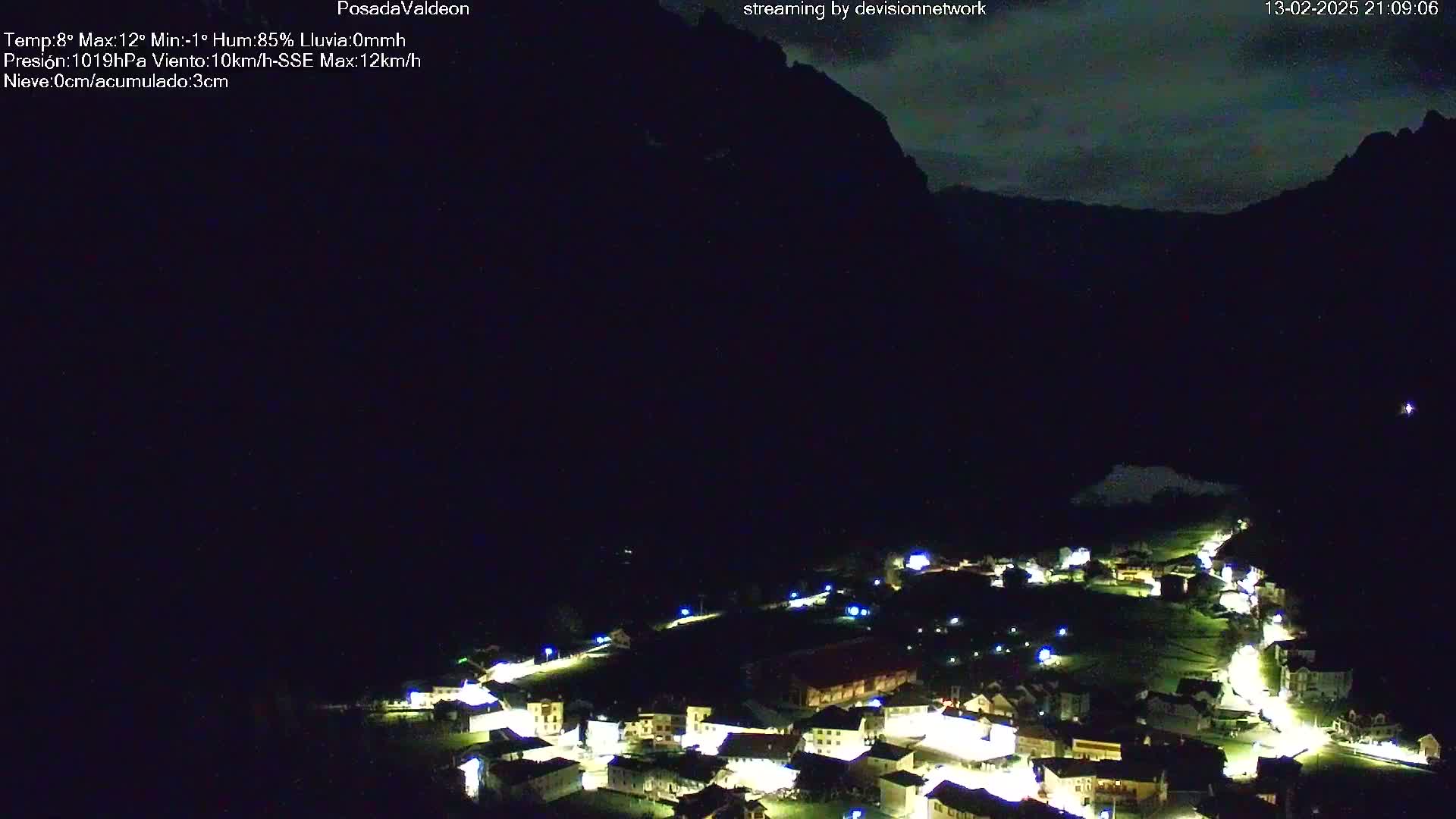 A nighttime view of a village nestled in a valley below a dark mountain range under a mostly clear sky.