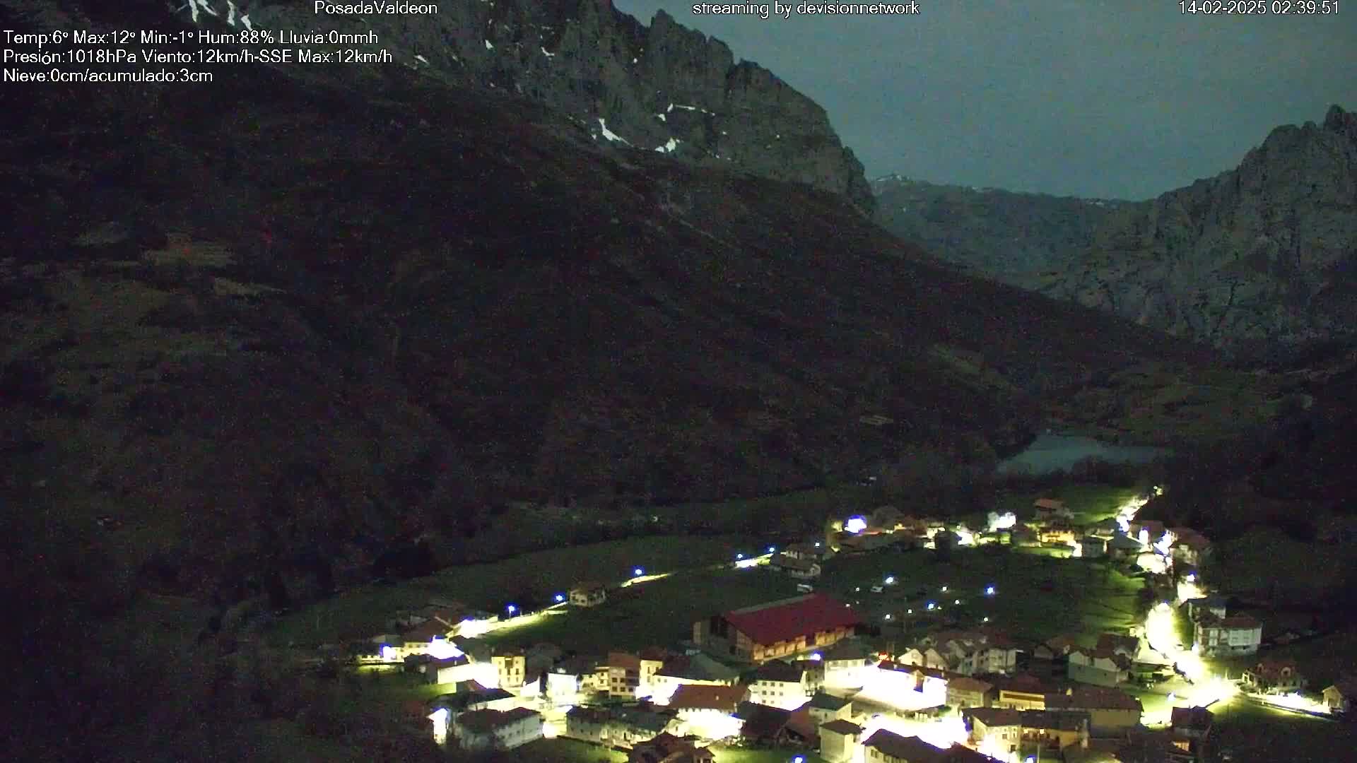 A small, illuminated village nestled in a dark valley between towering, snow-dusted mountains under a twilight sky.