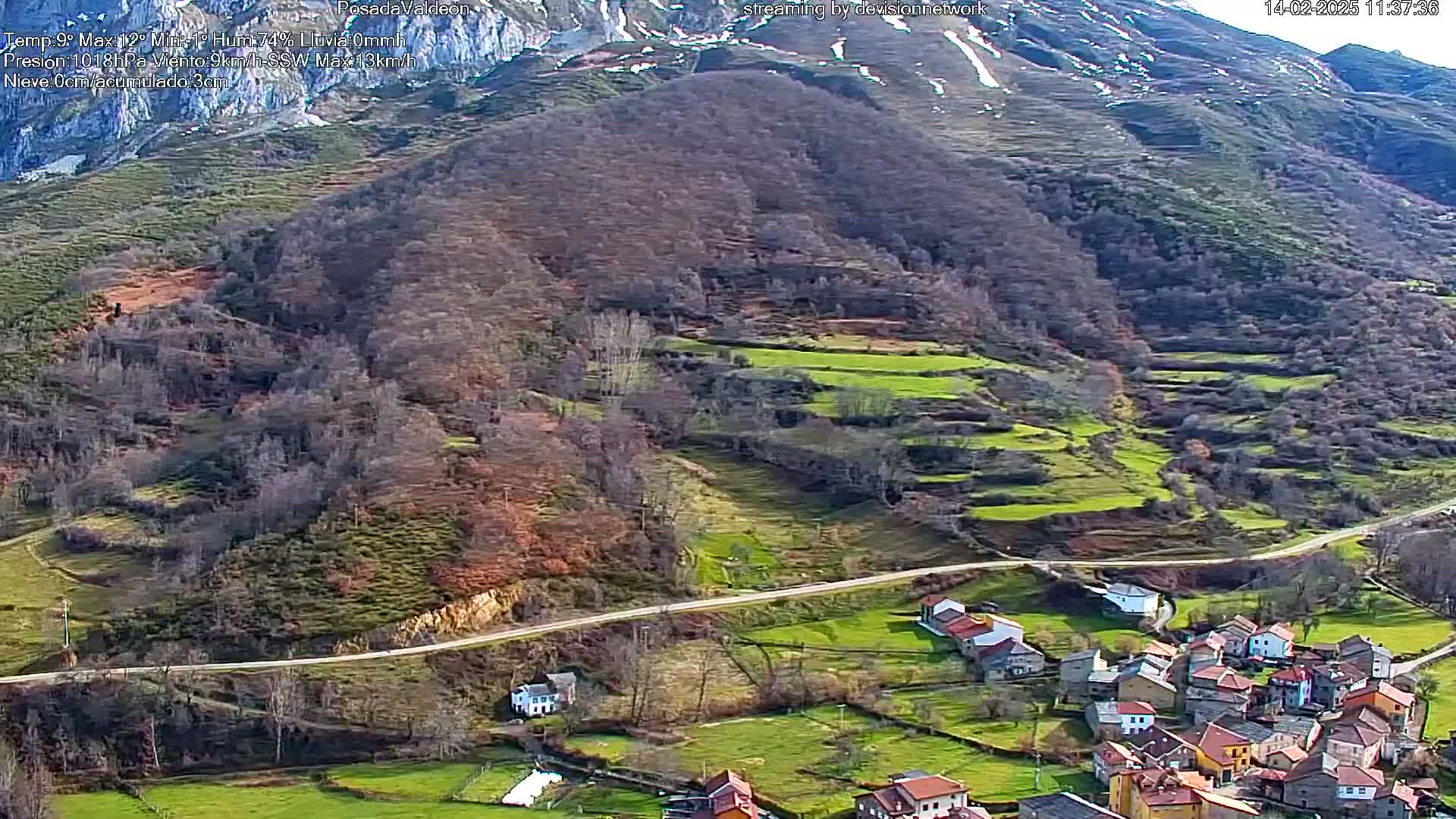 A mountainous landscape with a small village nestled in a valley, partially snow-capped mountains in the background, and green pastures interspersed with bare trees under a clear sky.