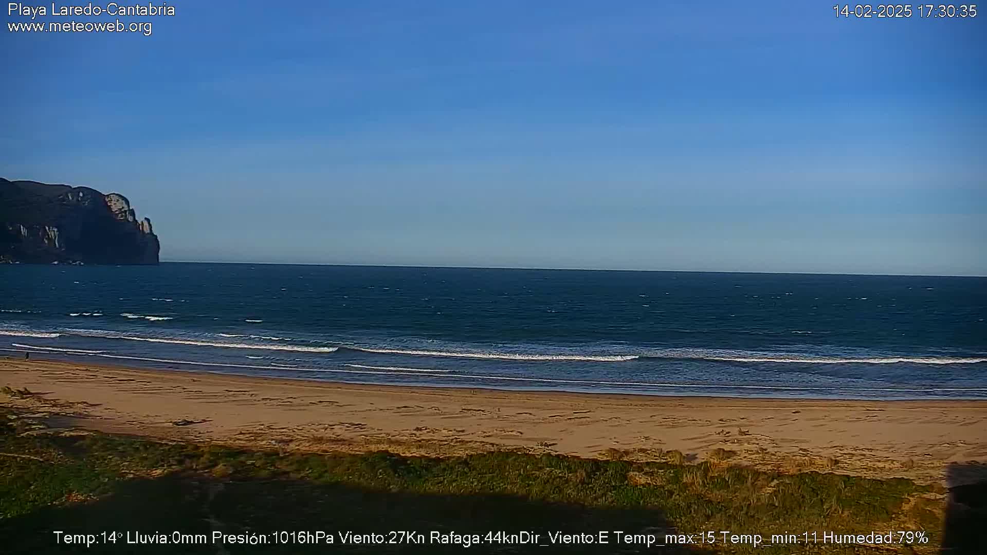 A sandy beach with gentle waves laps the shore under a clear blue sky, a dark cliff visible in the distance.