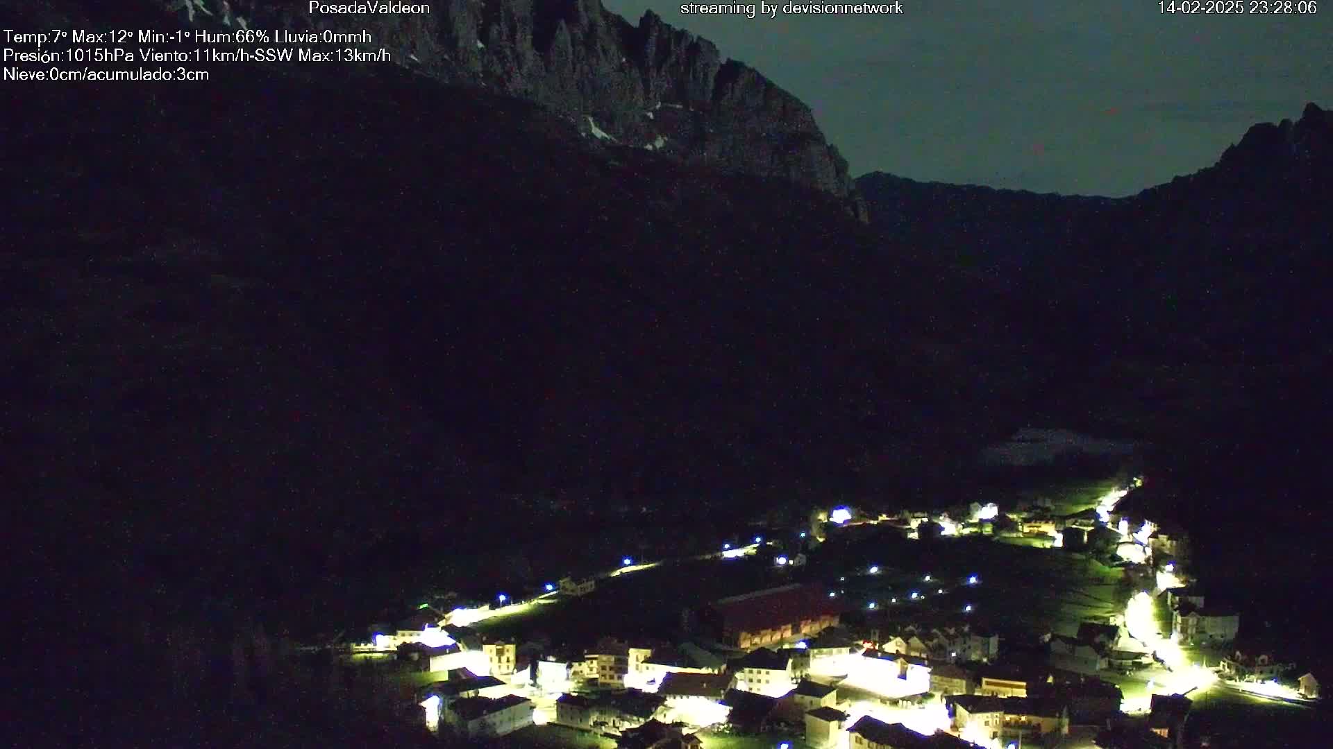 A nighttime view of a small village nestled in a valley, illuminated by lights, with dark mountains rising in the background under a mostly clear sky.