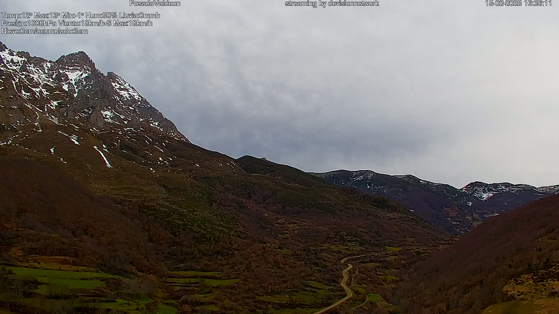 A winding road cuts through a valley nestled between snow-dusted mountains under a cloudy sky.