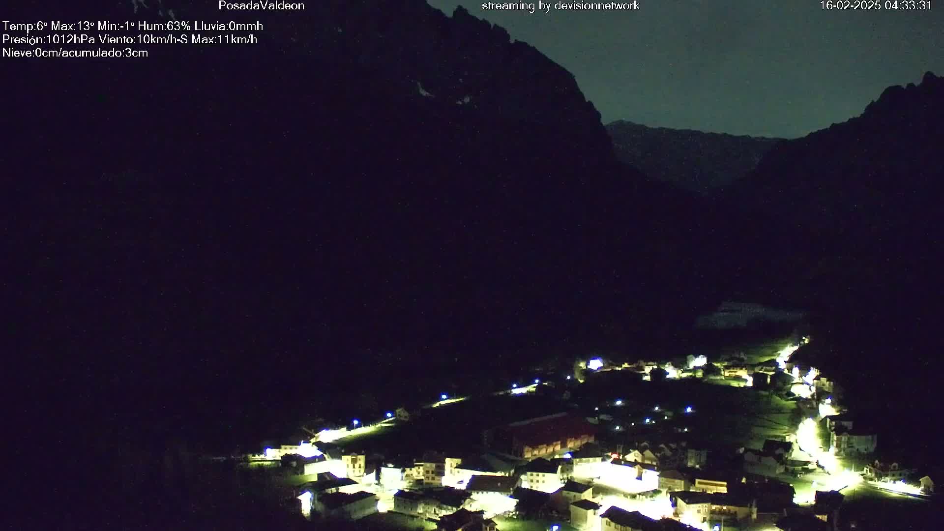 A nighttime, aerial view of a small village nestled in a valley, illuminated by streetlights, with dark mountains looming in the background under a clear, dark sky.
