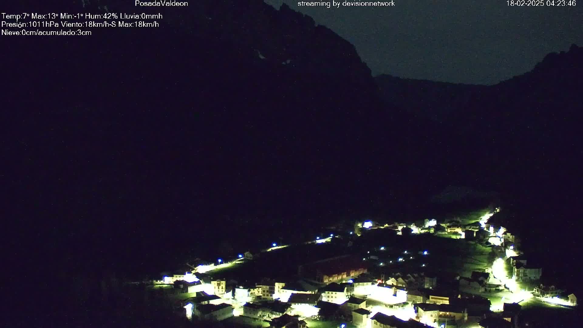 At night, a small village with illuminated buildings nestles below dark mountains under a clear, dark sky.