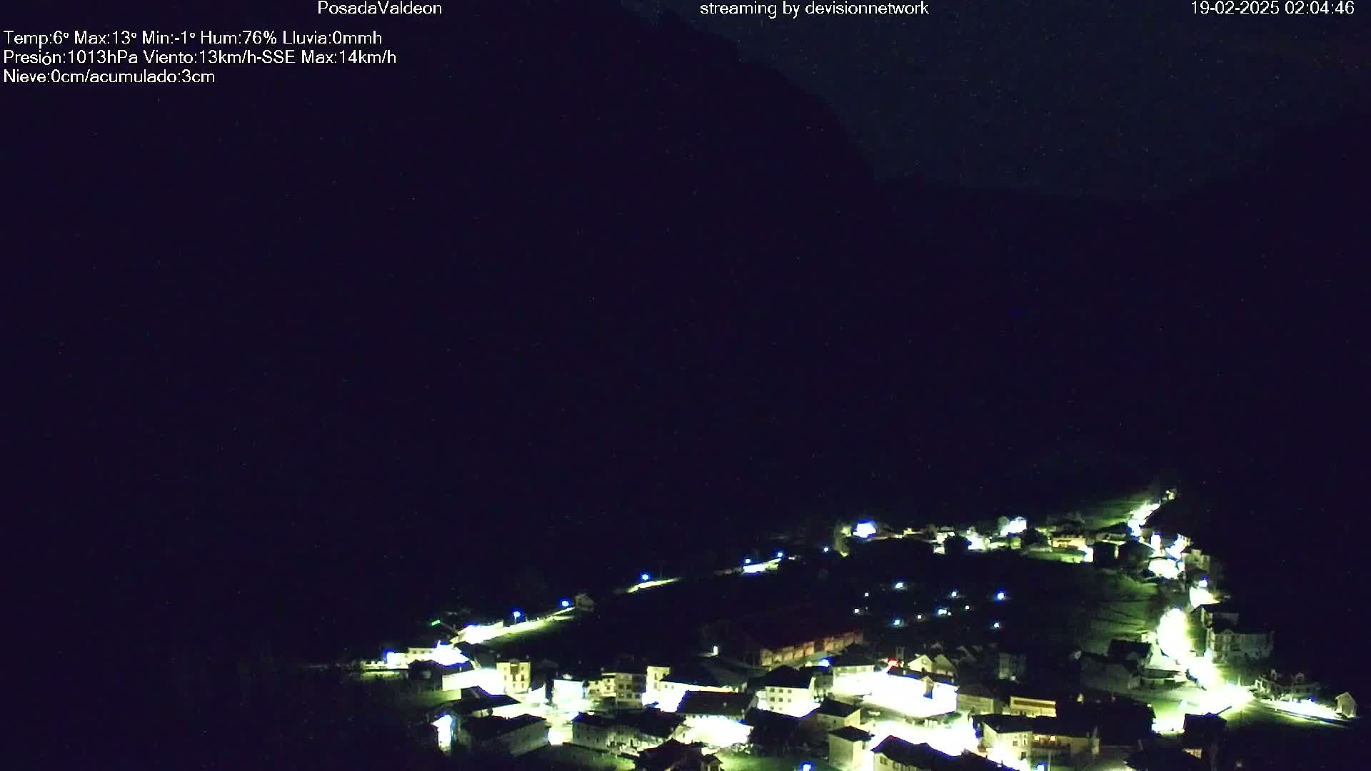 A small village at night, illuminated by streetlights, is nestled in a dark valley under a clear, starlit sky.