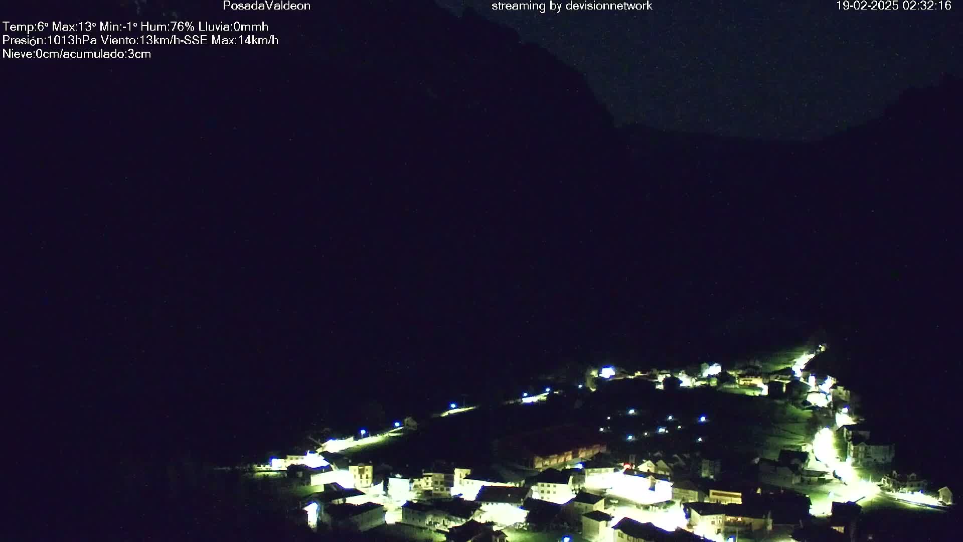 A small village illuminated at night is nestled below a dark mountain under a clear, starlit sky.