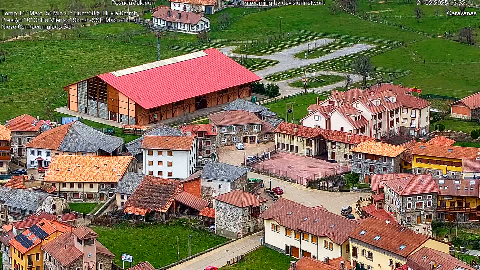 An aerial view of a village with red-tiled roofs and a large red barn, under partly cloudy skies.
