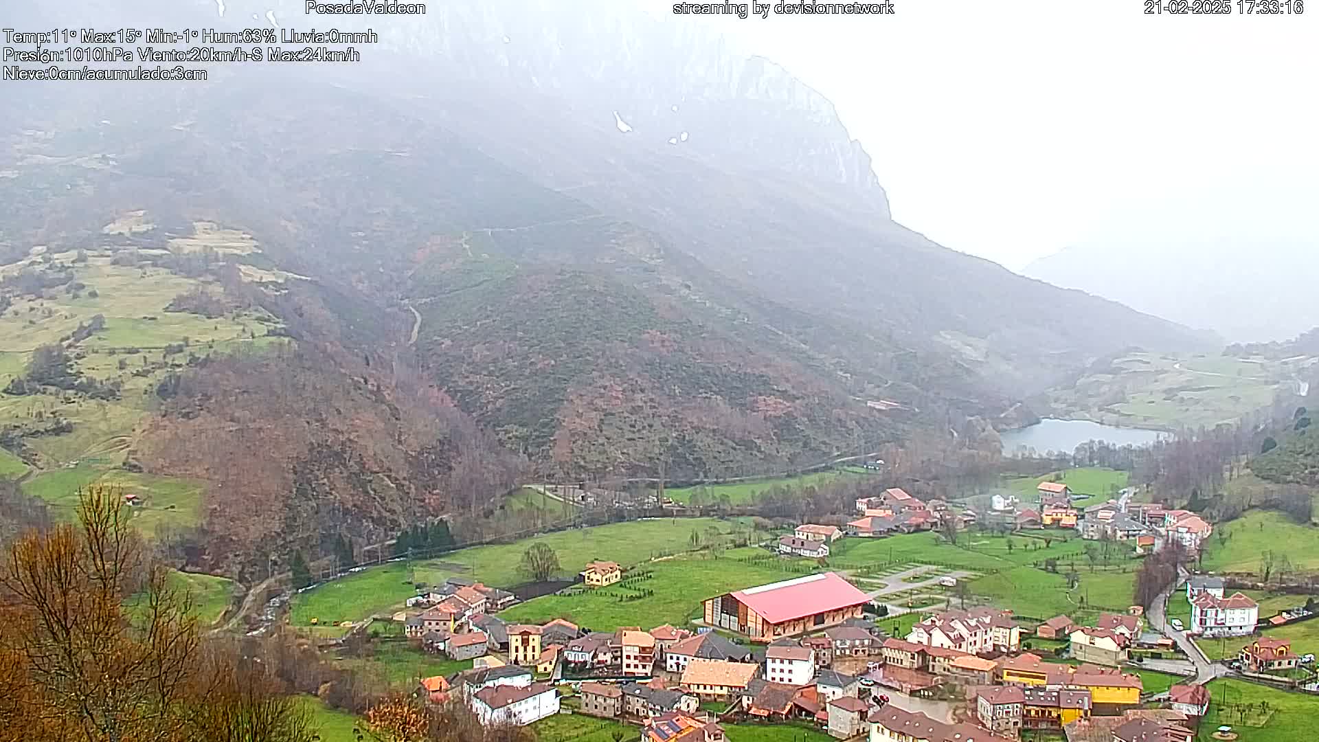 A small village nestled in a valley surrounded by mist-covered mountains and a lake, under overcast conditions.