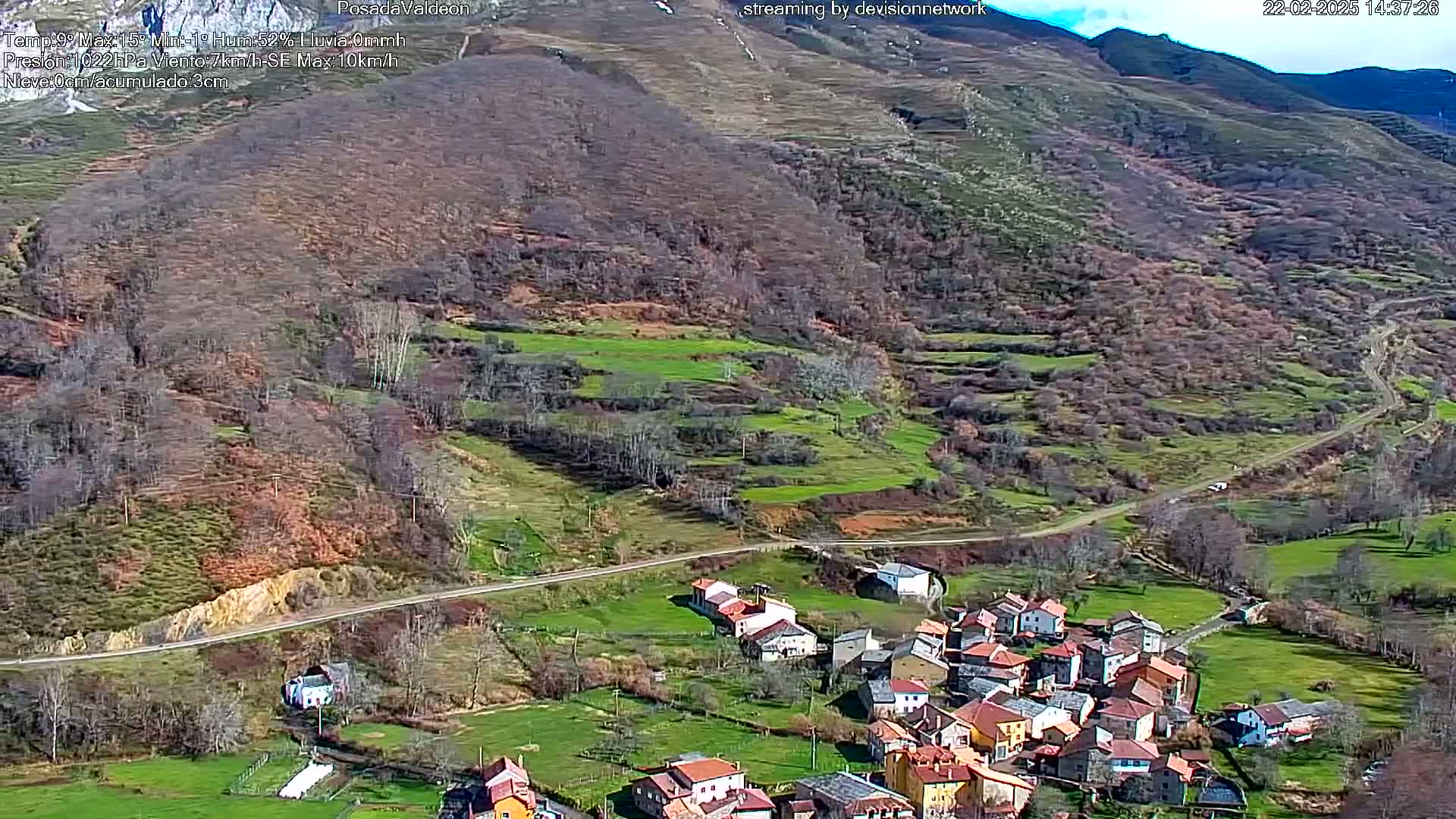 A small village nestled in a valley of green fields and brown, leafless trees, under a mostly clear sky.