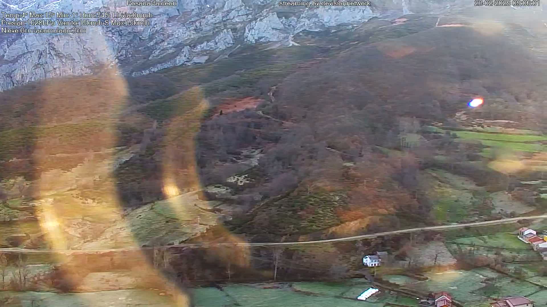 A high-angle, long shot of a mountainous landscape with a valley below, showing a road winding through the valley floor, and some small buildings nestled amongst the frost-covered fields, under somewhat hazy conditions.