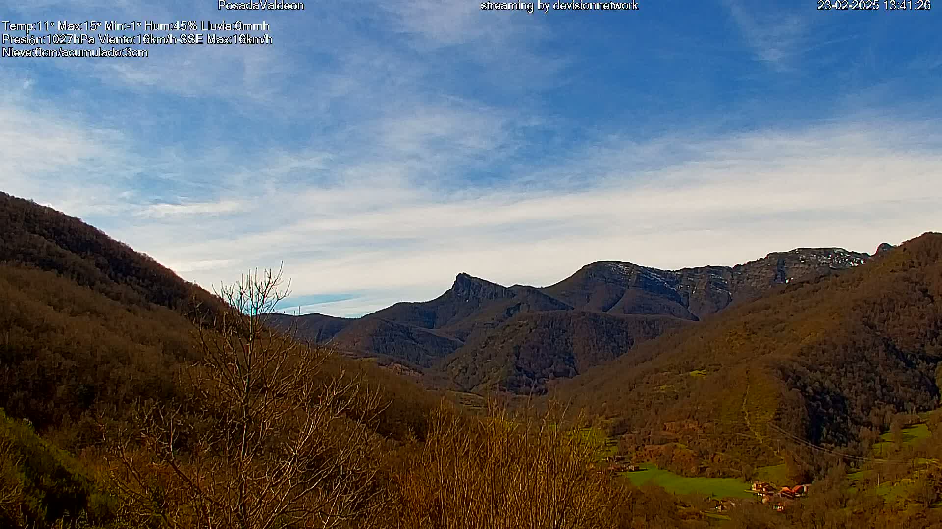 A mostly clear blue sky overlooks a valley of brown, leafless trees and distant mountains with patches of snow.