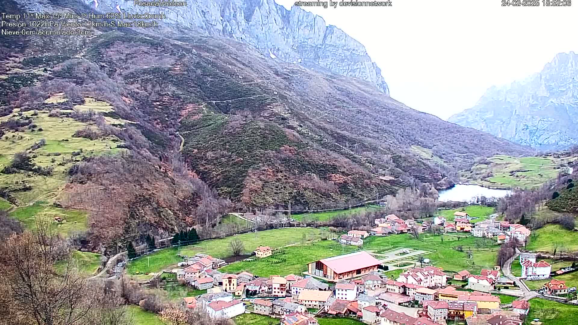 A small village nestled in a valley between two mountains, under a mostly cloudy sky, features a lake and green fields.