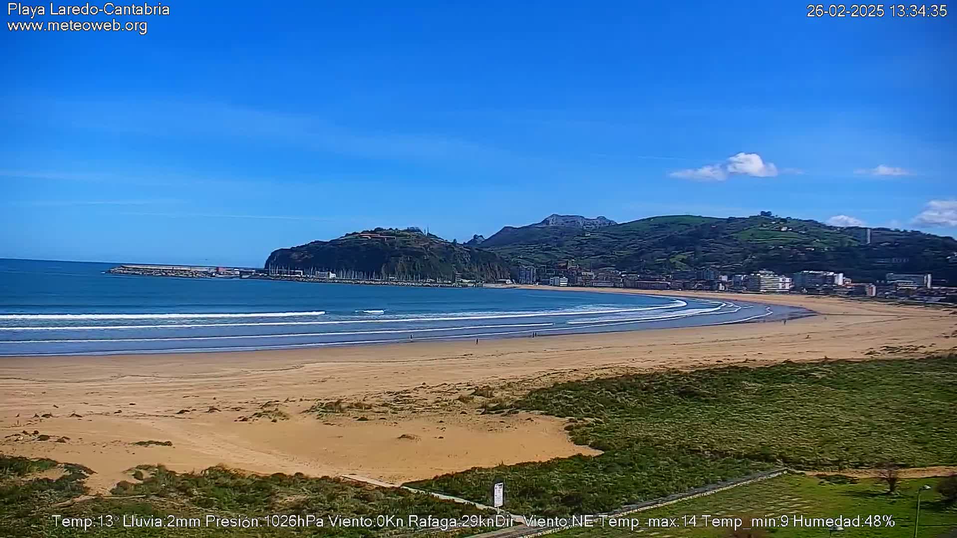 A sandy beach with gentle waves laps the shore under a clear blue sky, backed by a town nestled against a verdant hill.