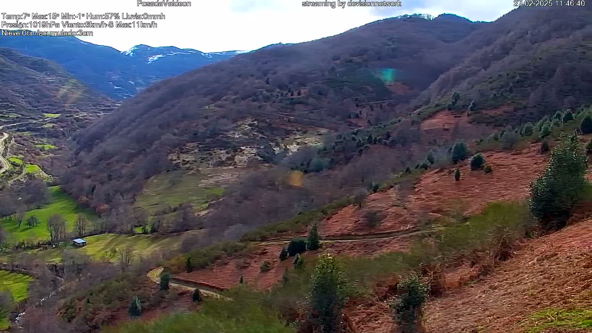 A valley with sparsely wooded hillsides, some showing reddish-brown patches of vegetation, under a partly cloudy sky.