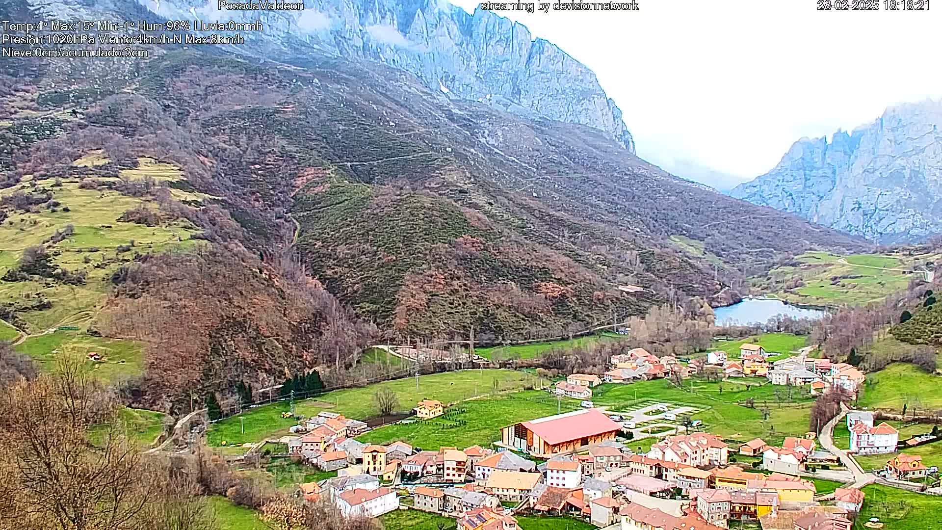 A small village nestled in a valley between towering mountains on an overcast day, with a small lake visible in the valley.