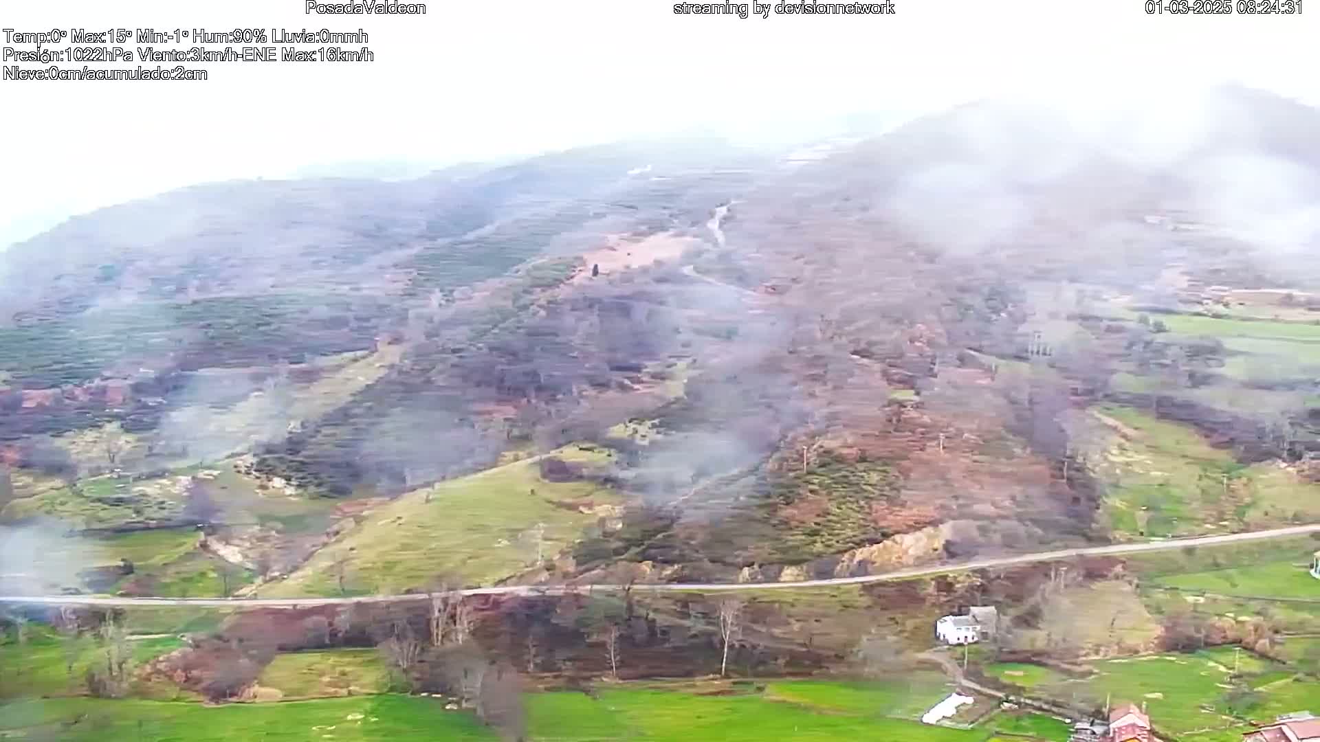 A high-angle view of a hilly landscape with some smoke rising from a hillside, under hazy, foggy conditions.