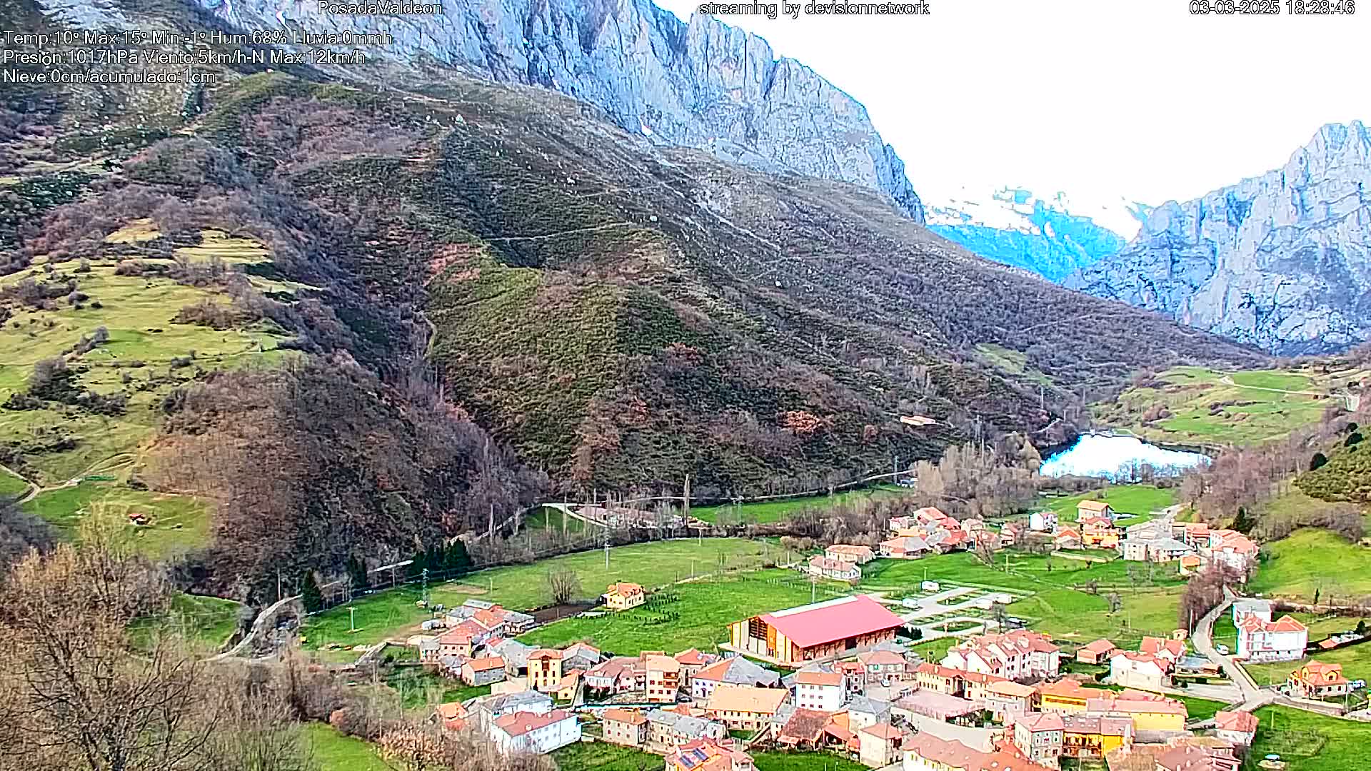 A small village nestled in a verdant valley between towering, snow-dusted mountains under a mostly clear sky.