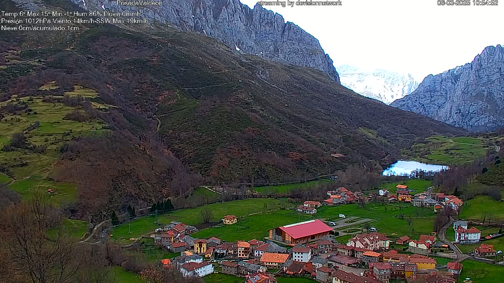 A small village nestled in a valley between towering mountains, with a small lake nearby, under mostly cloudy skies.