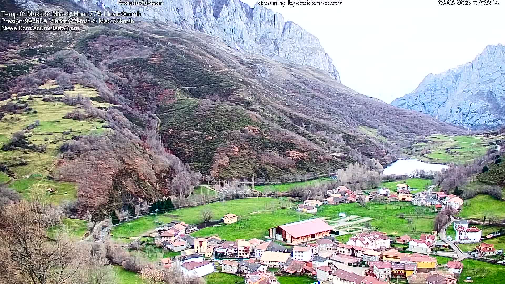 A small village nestled in a valley between towering mountains, under a mostly cloudy sky.
