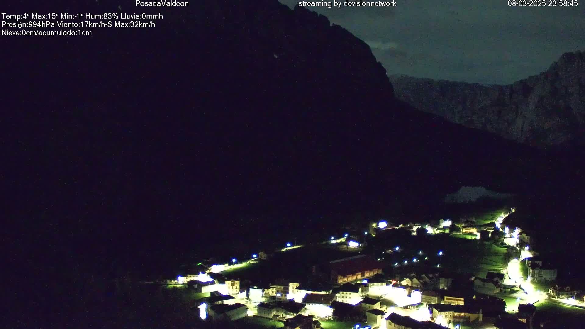A nighttime view of a small village nestled in a valley, illuminated by lights, with a dark mountain looming in the background under a clear, dark sky.