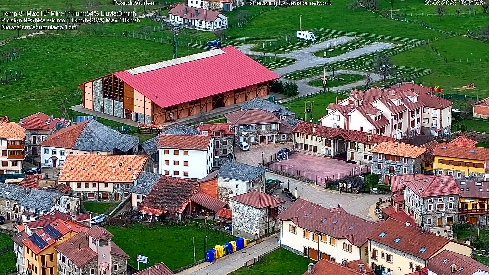 An aerial view of a small village with red-roofed buildings, a large red building, and a paved area, under partly cloudy skies.