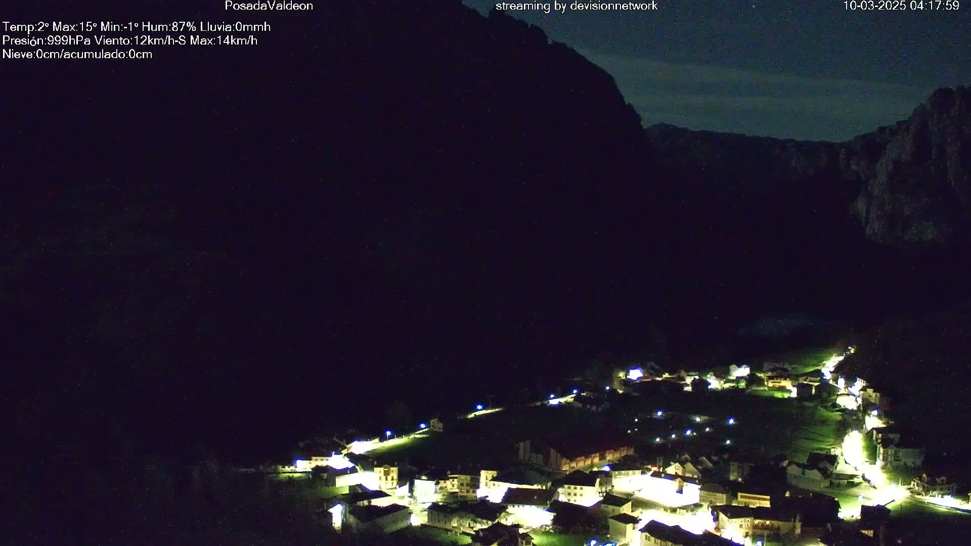 A small village illuminated at night is nestled below a dark mountain under a mostly clear night sky.