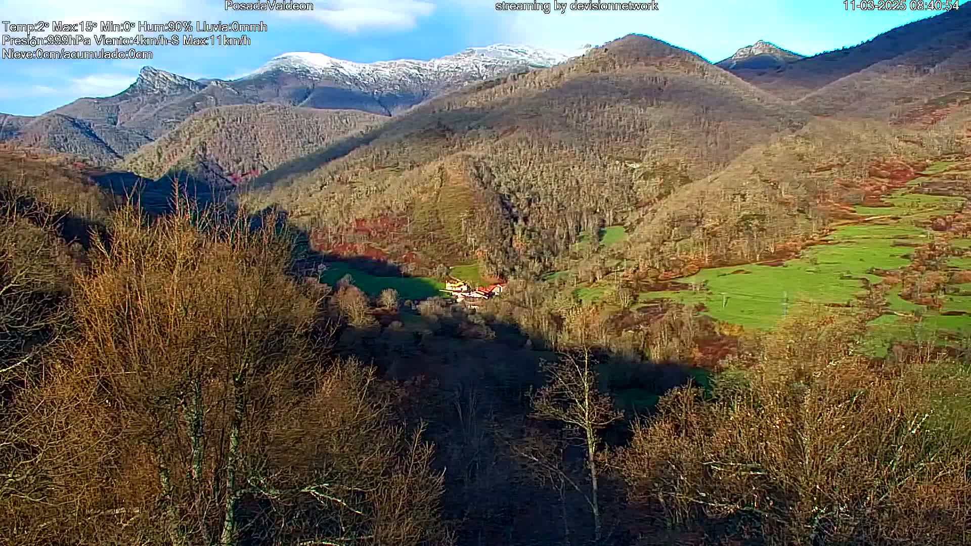 A sunny, partly cloudy view of a valley nestled among snow-capped mountains, with sparse, mostly leafless trees and patches of green pastureland.