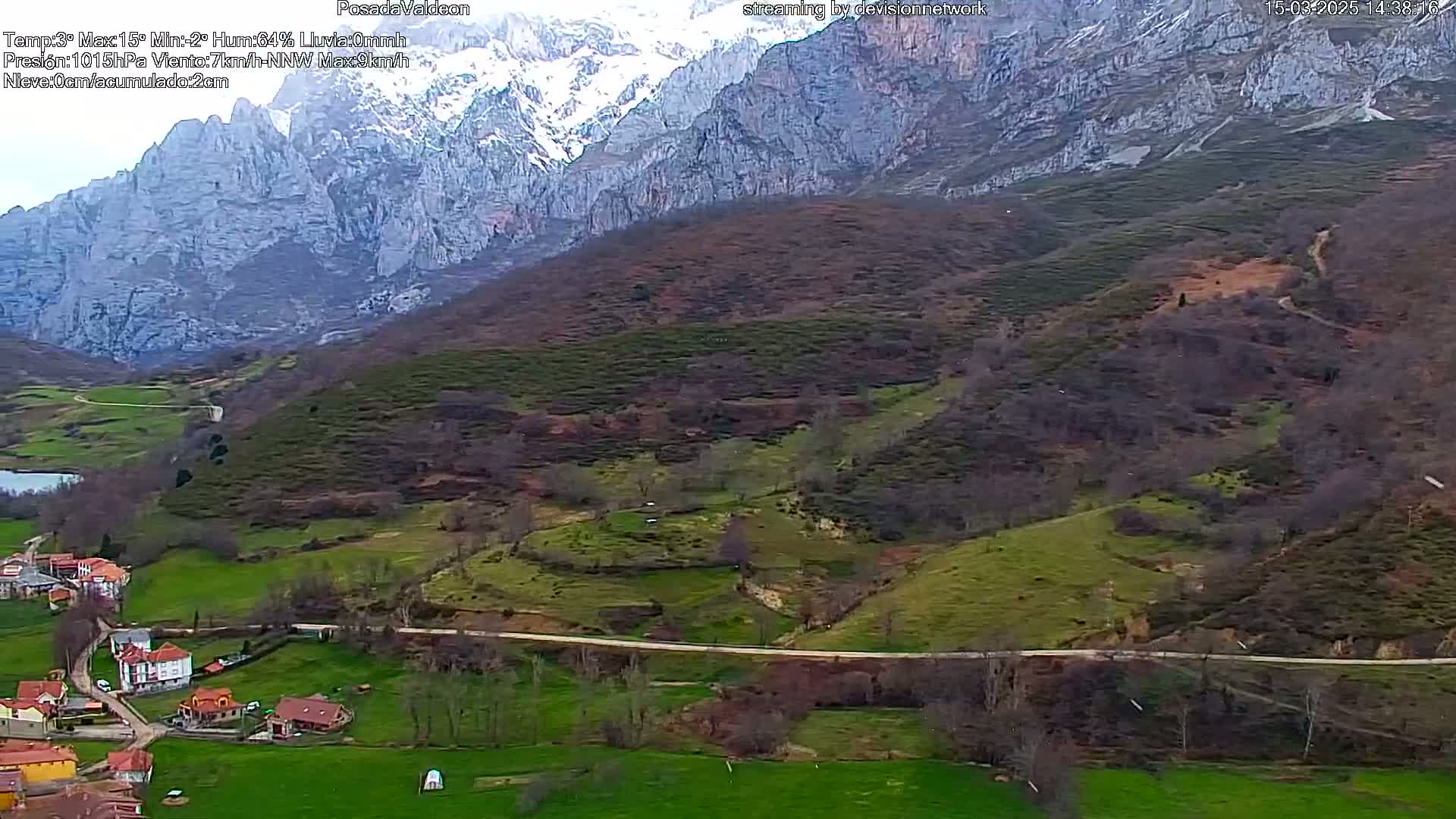 A high-angle view shows a valley town nestled below snow-capped mountains, with a road winding through green and brown hills under an overcast sky.