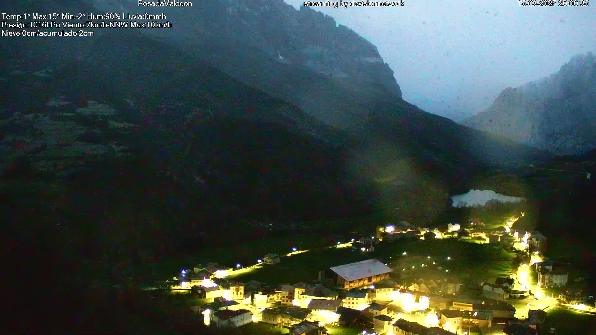 A nighttime view of a small, illuminated village nestled in a valley between dark mountains under a cloudy, rainy sky.