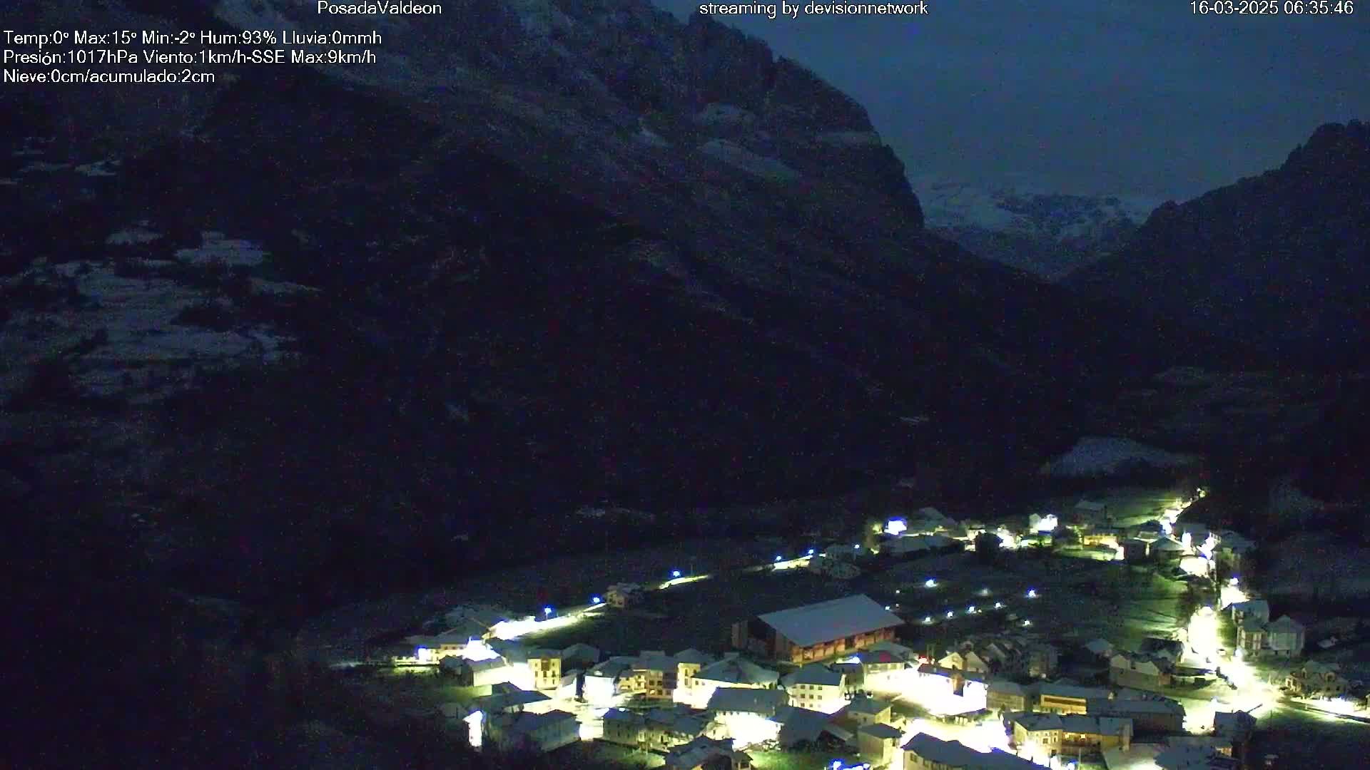 A small, snow-covered village nestled in a dark valley under a night sky, illuminated by its streetlights and with mountains in the background.
