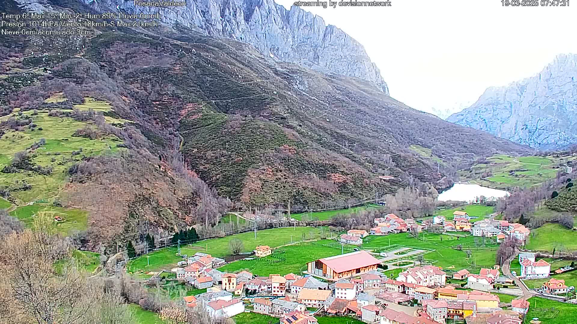 A small village nestled in a valley between mountains, with a small lake nearby, under an overcast sky.