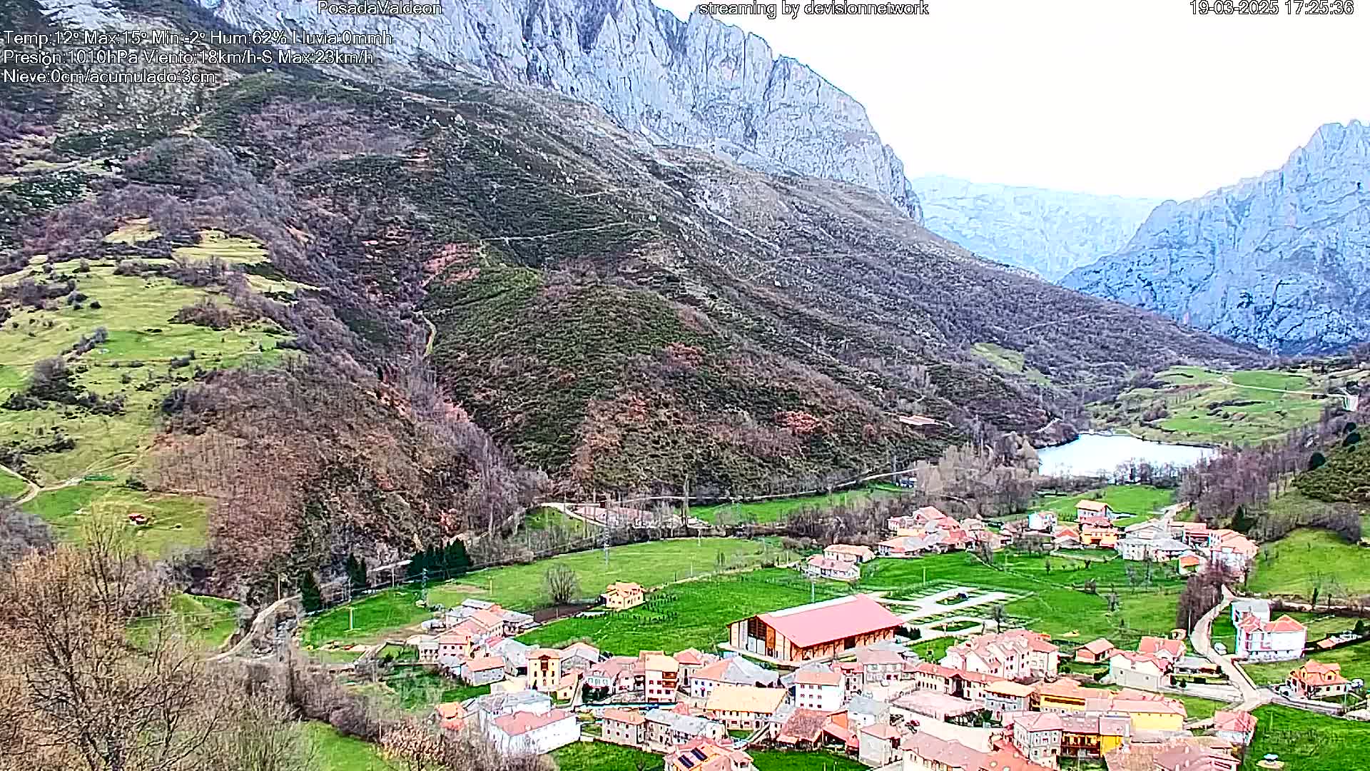 A small village nestled in a verdant valley between imposing mountains, under a partly cloudy sky.