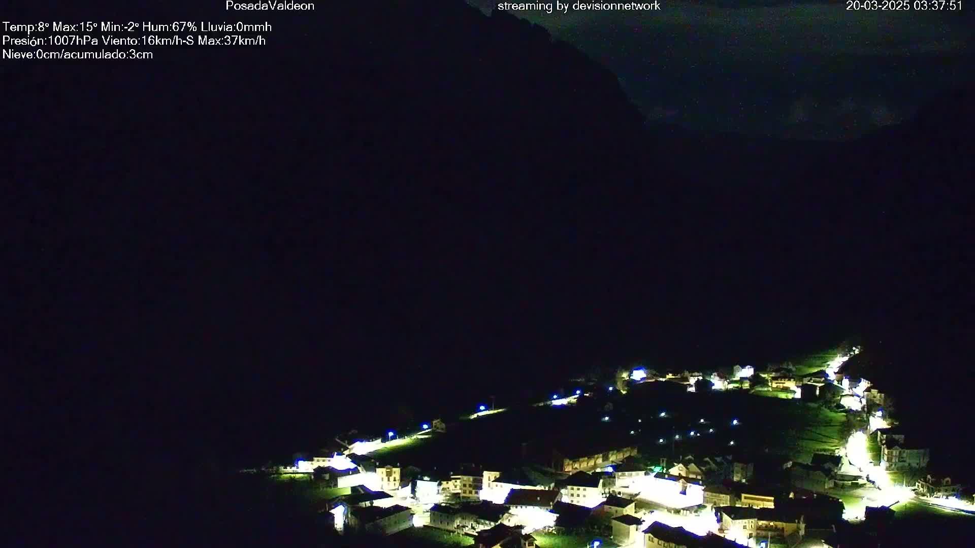 At night, a small village nestled in a valley is illuminated by lights, with dark mountains rising in the background under a clear, dark sky.