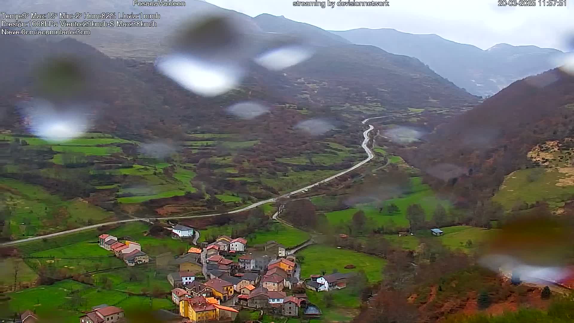 A rain-covered view shows a small village nestled in a valley between green and brown hills, with a winding road leading away.